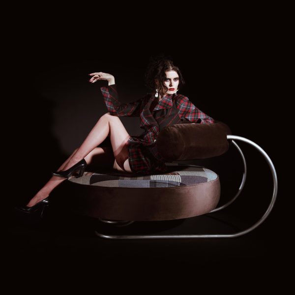 A person is laid across a large circular seat with a brown, curved, cylindrical back stop that is held up by a steel frame. The circular seat has patchwork upholstery.
