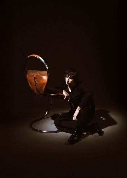 Image of a person sitting on the floor and leaning on a chair made from tanned leather that has been wrapped around a stainless steel frame. The leather has laced detailing resembling a corset. 