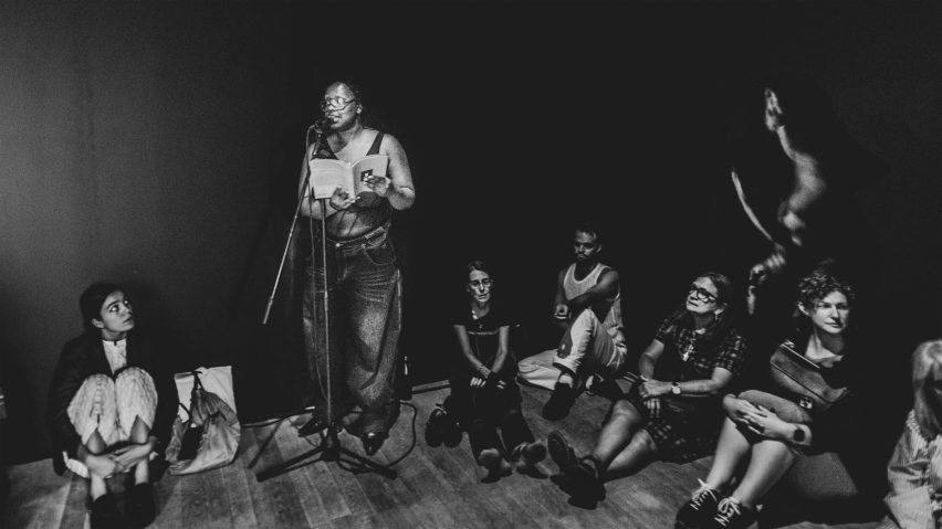 Photo of person using a microphone surrounded by other people sat down