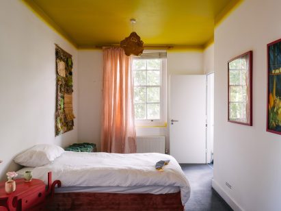 Photo of the "whimsical" bedroom at the UAL Atterbury Flat, featuring a chartreuse-coloured ceiling with a neon gradient effect as it merges into white walls, soft pink curtains, a shaggy textile work on one wall and a bedside table in the foreground that looks like an abstracted alien character