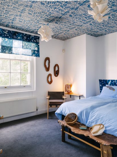 Photo of the "calm" bedroom at the UAL Atterbury Flat, with white walls, lots of wood and natural materials like paper ceiling lights, a cyanotype printed window dressing with a botanical motif, and a wallpapered ceiling with a blue-and-cream River Thames shoreline motif