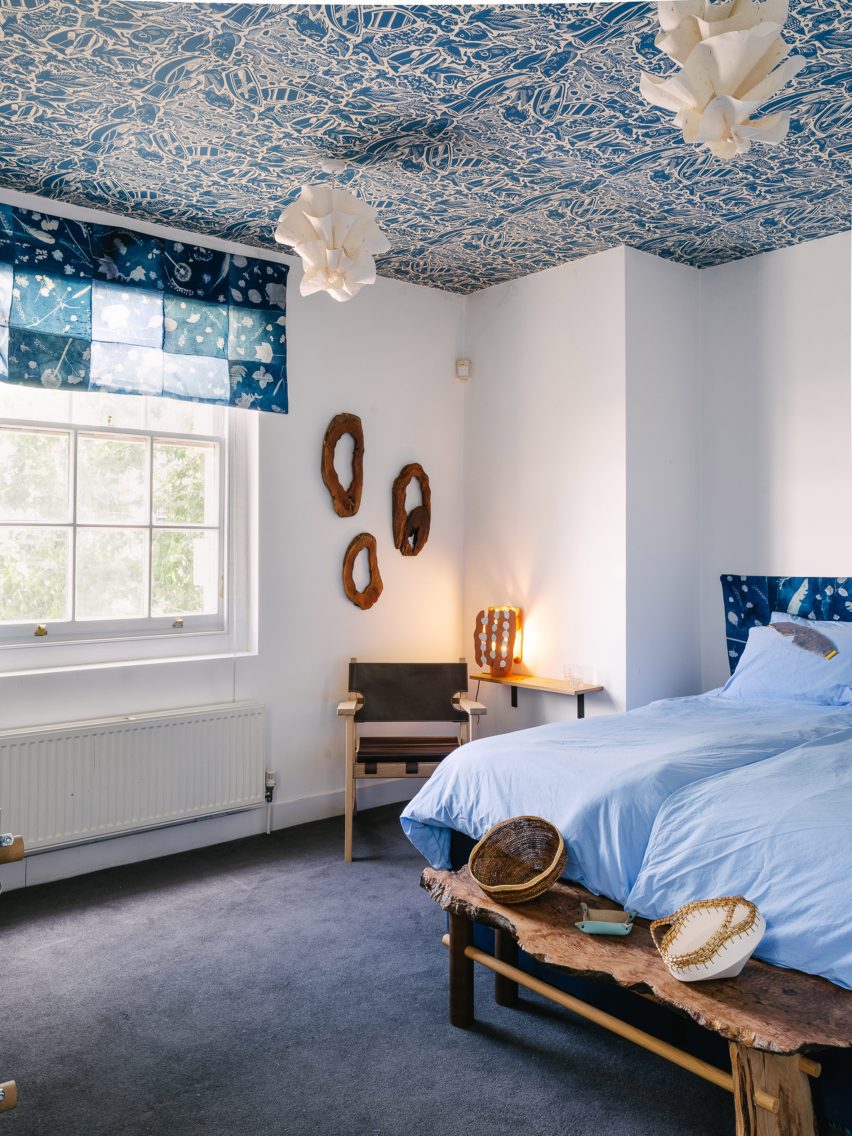 Photo of the "calm" bedroom at the UAL Atterbury Flat, with white walls, lots of wood and natural materials like paper ceiling lights, a cyanotype printed window dressing with a botanical motif, and a wallpapered ceiling with a blue-and-cream River Thames shoreline motif