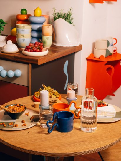 Close-up photo of the small circular wooden dining table at the UAL Atterbury Flat laden with ceramic dishes and food, with a colourful kitchen viewable in the background 