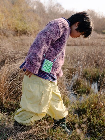 Photo of a child walking through grassy marshes and peering into the water while wearing wellington boots and their Freckle phone on a lanyard around their neck