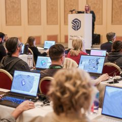 Attendees sitting at tables using laptops during a SketchUp workshop, with a presenter standing at a lectern at the front of the room