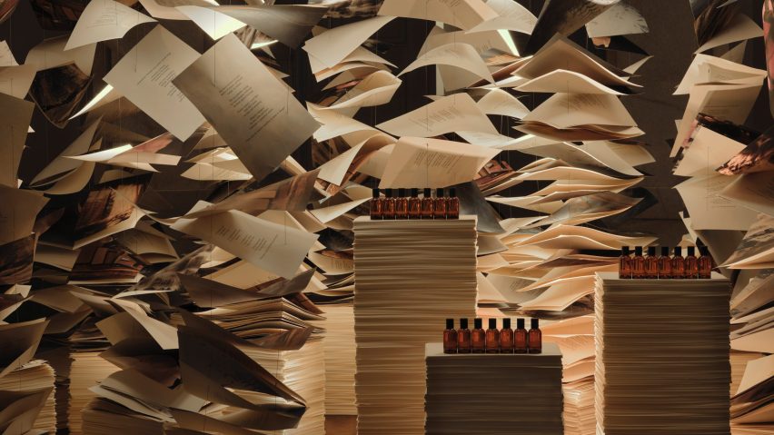 Aesop fragrance bottles displayed on stacks of books beneath suspended paper sheets